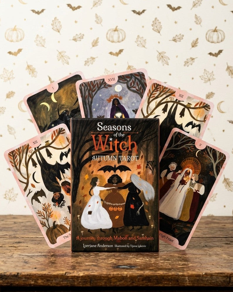 A boxed tarot deck titled "Seasons of the Witch" stands on a rustic wooden table with several illustrated tarot cards fanned out behind it. The artwork features autumn and witch motifs, including pumpkins, bats, trees, and figures around a cauldron.