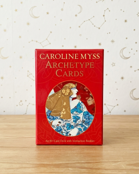 Caroline Myss Archetype Cards deck box standing upright on a light wood surface against a white wall with gold celestial patterns.
