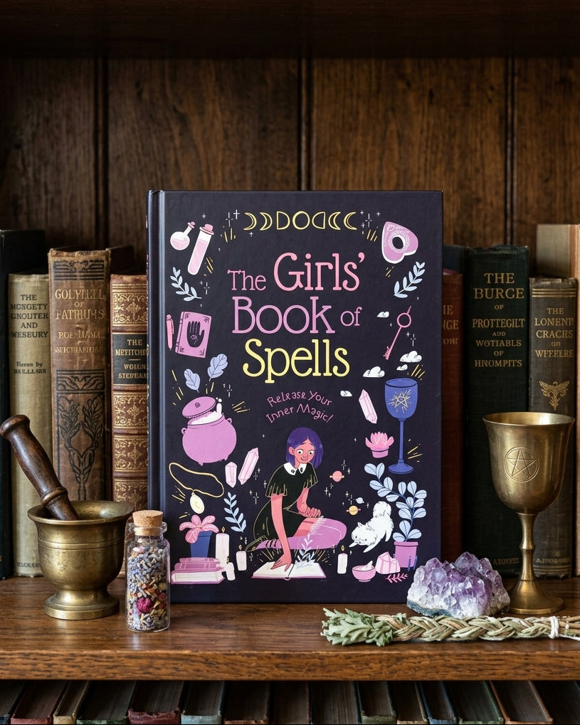 The Girls' Book of Spells standing upright on a wooden shelf, surrounded by old books, a brass mortar and pestle, a small bottle, a crystal ball, and a silver wand.