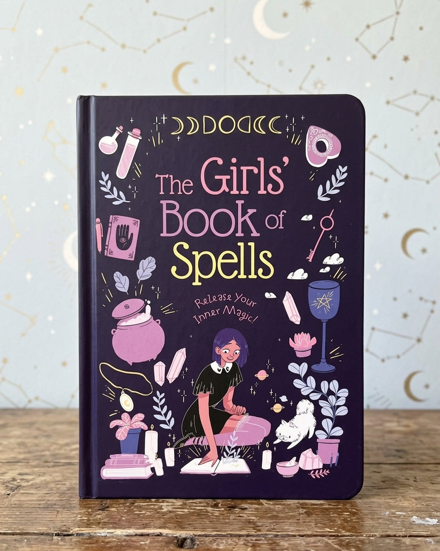The Girls' Book of Spells notebook standing upright on a rustic wooden table against a light blue wall with celestial patterns. The notebook cover is dark purple with whimsical illustrations of magical items and a girl casting a spell.