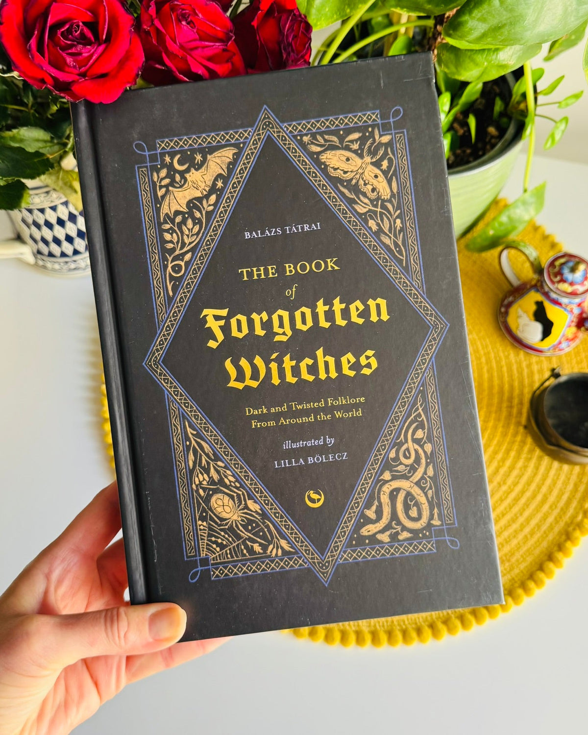 A hand holds a hardcover book titled "The Book of Forgotten Witches" by Balaji Jaybai. The book cover is dark gray with ornate gold and blue decorative borders and gold lettering.
