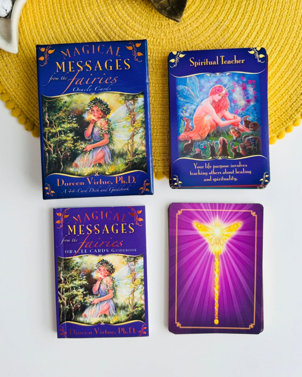 A boxed card deck and two sample cards from the 'Magical Messages from the Fairies' oracle deck are arranged on a white surface with a yellow woven placemat and decorative props around them. The box and cards feature blue borders and fantasy illustrations, including a glowing wand on the back of a card.