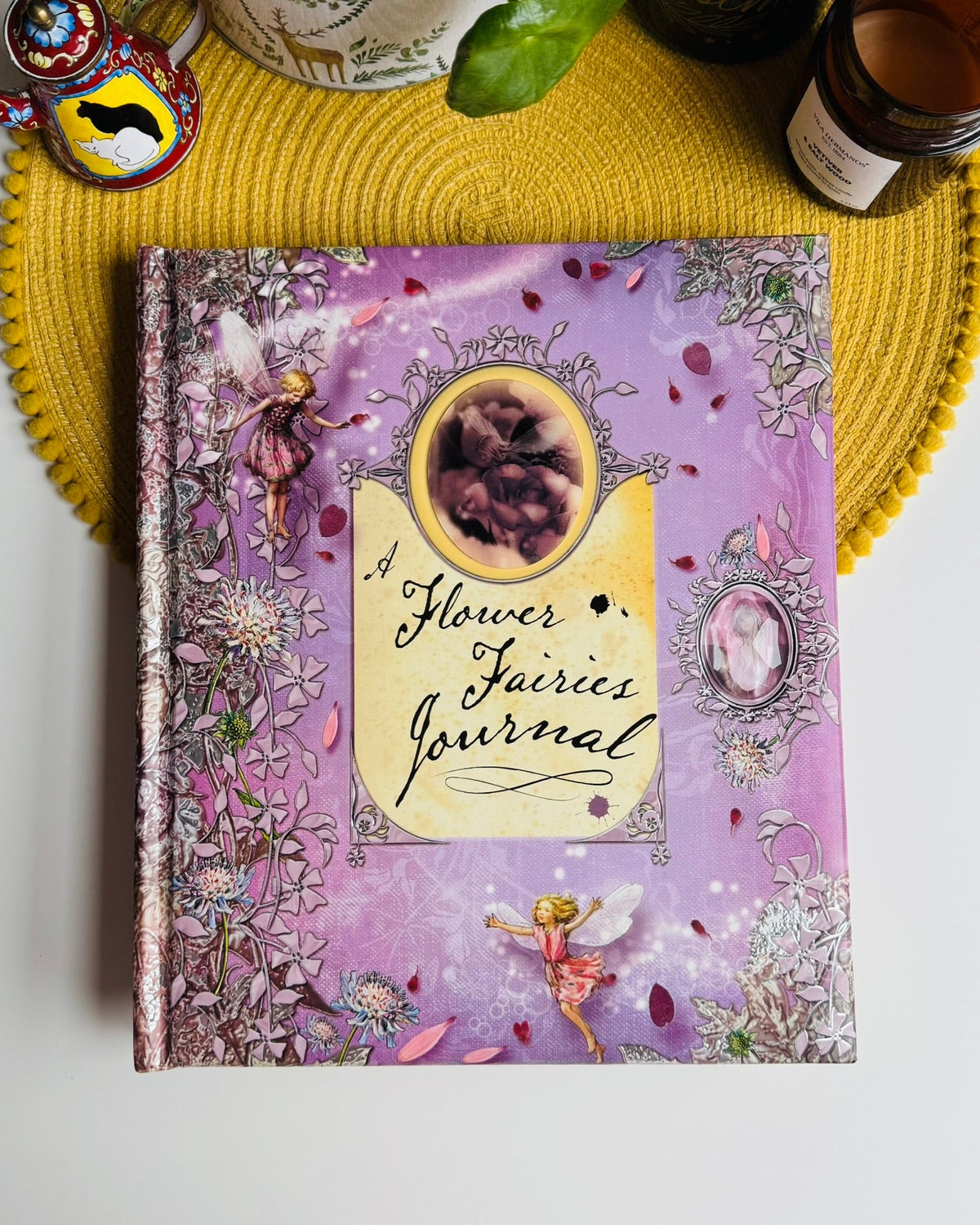 A decorative hardcover journal titled "A Flower Fairies Journal" lies on a white surface, styled with a yellow woven placemat and surrounding props including a small teapot, candle, and ceramic dish.