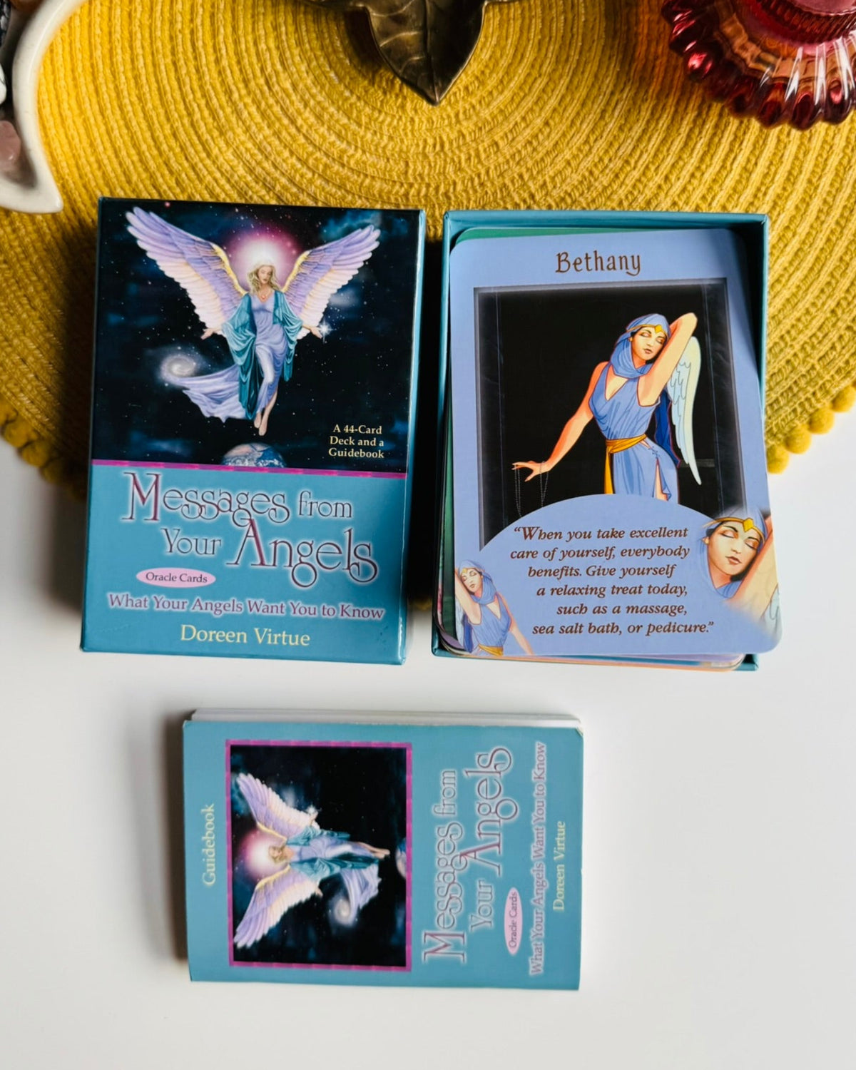 A boxed card deck and two individual cards from 'Messages from Your Angels' by Dorsey Virtue are arranged on a white surface with a yellow woven placemat and decorative props around them.