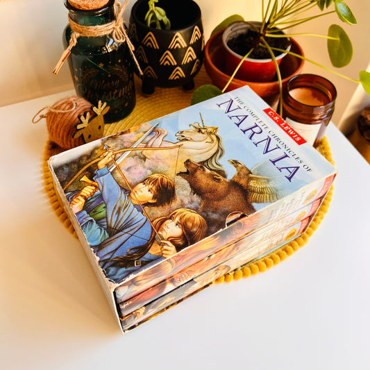 A boxed set of Narnia books styled on a white tabletop with plants, a candle, and decorative jars in the background. The box cover shows the title 'NARNIA' and 'COMPLETE COLLECTION VOLUME 1' with illustrated characters and animals.