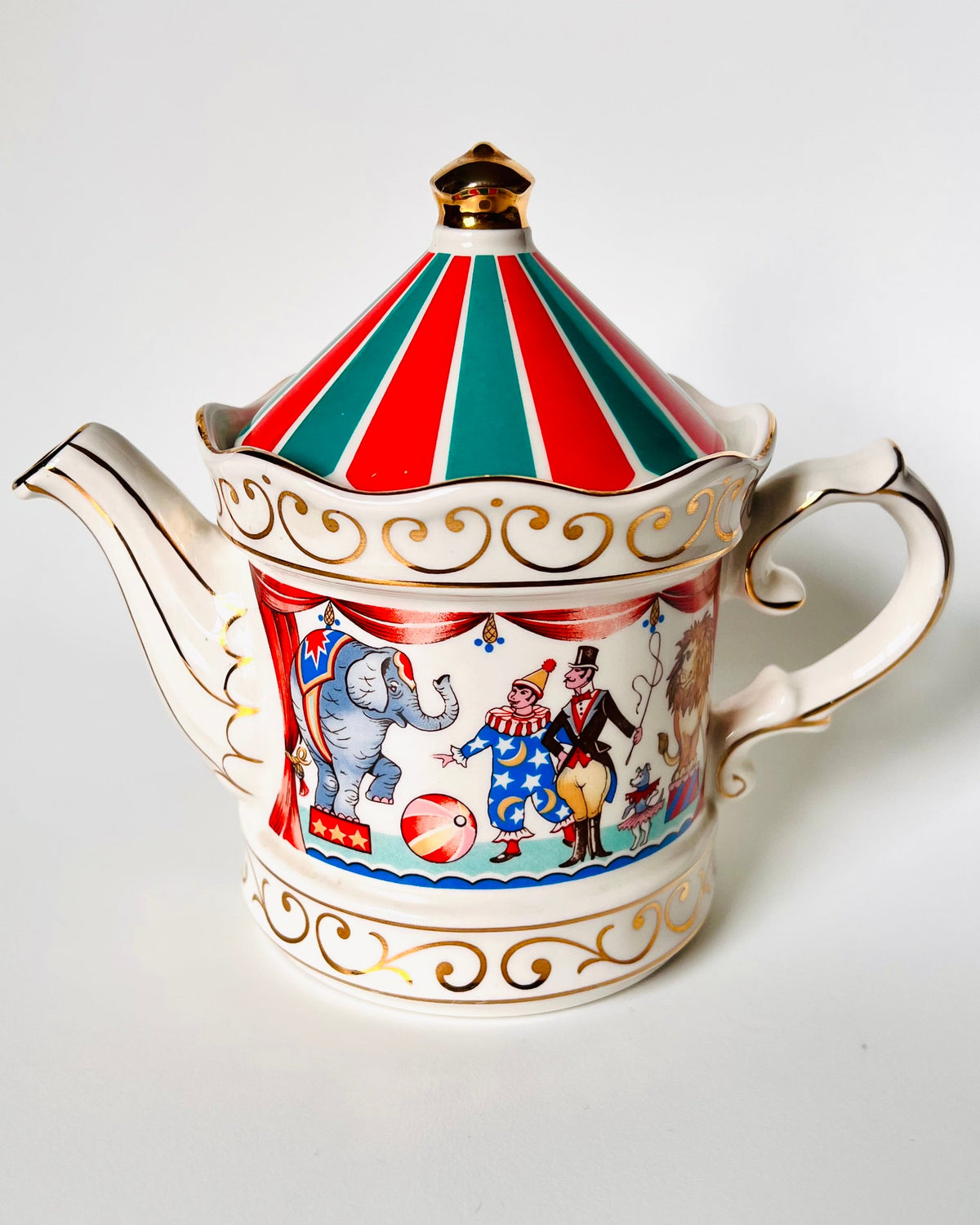 A vintage porcelain teapot with a red and green striped lid, gold trim, and a colorful circus-themed illustration on the body, set against a plain light gray background.