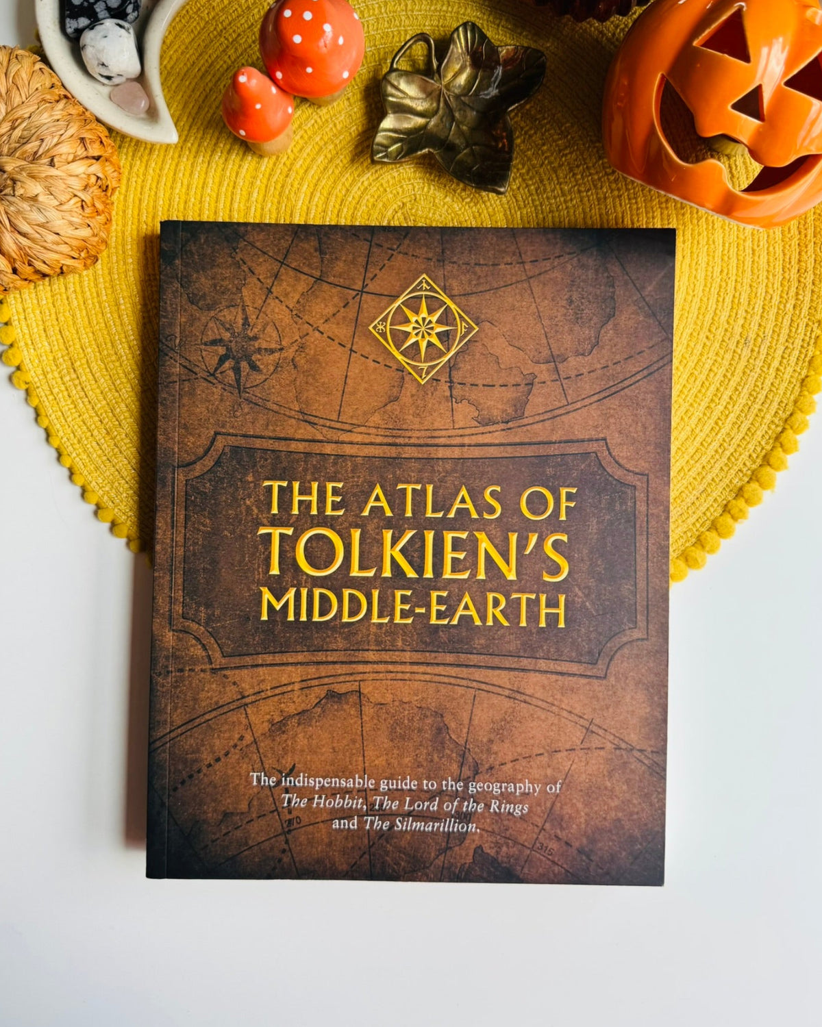 A book titled "The Atlas of Tolkien's Middle-earth" lies on a yellow woven placemat, surrounded by autumn props including a carved pumpkin, small pumpkins, a decorative leaf, and stones.