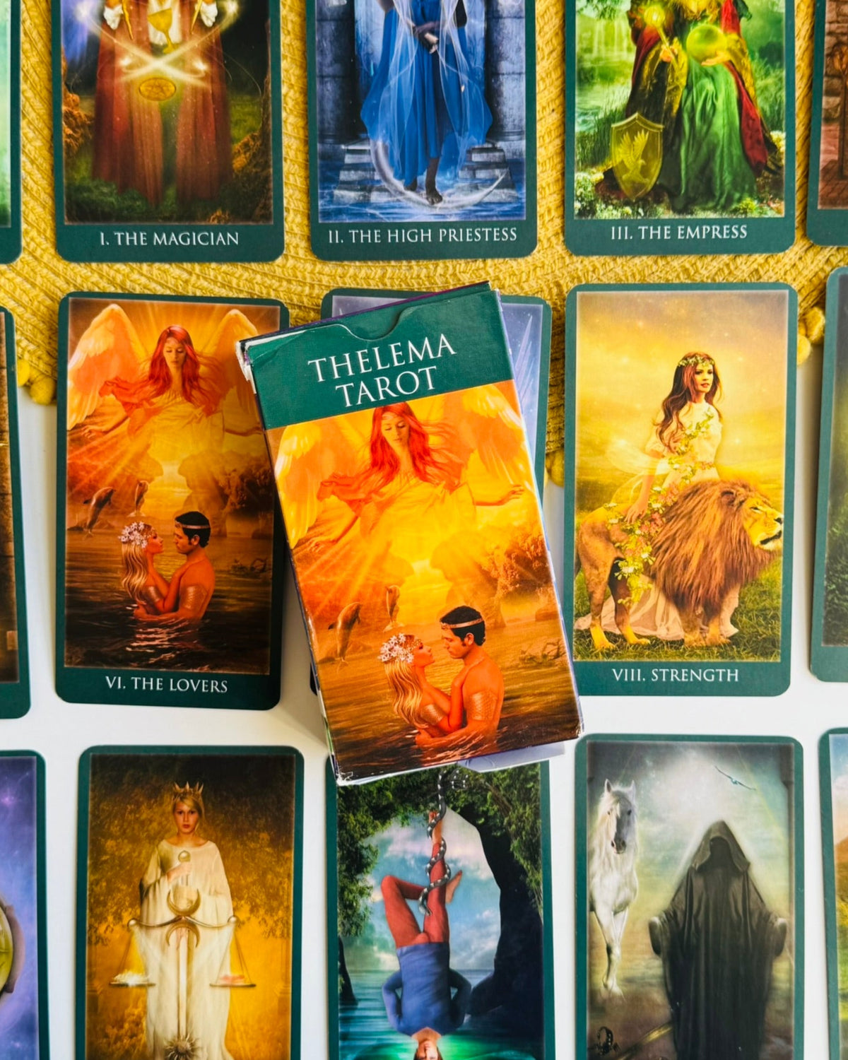 A Thelema Tarot deck with its box placed on top of several illustrated cards, arranged on a white surface with a gold rope border. The front of the box reads 'THELEMA TAROT' and shows a fiery orange-toned illustration.