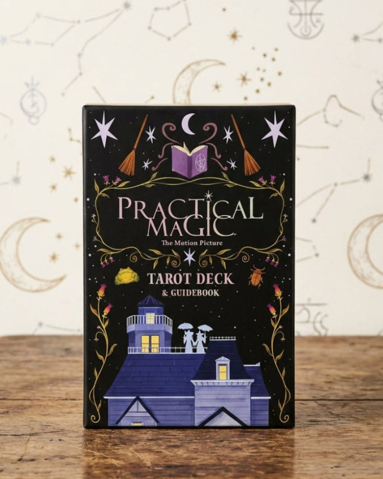 A boxed tarot deck product standing upright on a rustic wooden table against a cream wallpaper background with moon and constellations. The box is black with decorative gold and purple artwork and the title 'Practical Magic Tarot Deck' visible.