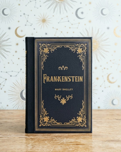 A hardcover book of Frankenstein by Mary Shelley, featuring a dark navy blue cover with ornate gold foil detailing and lettering, standing upright on a light wooden surface against a patterned wallpaper background.