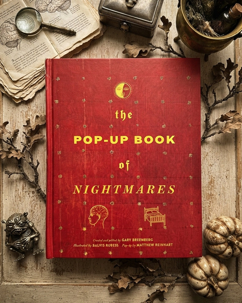 A red hardcover pop-up book titled 'The Pop-Up Book of Nightmares' is displayed in a styled flat-lay arrangement on a rustic wooden surface, surrounded by vintage books, a small metal box, bottles, dried leaves, and pumpkins.