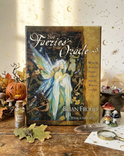 The 'Los Faeries Oracle' deck box by Brian Froud is displayed on a rustic wooden table surrounded by autumn-themed props including a small bottle of fairy dust, a carved pumpkin, leaves, and miniature mushrooms against a cream background with gold moon and star illustrations.