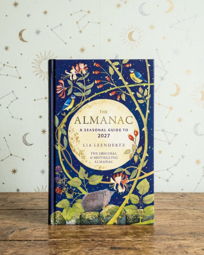 The Almanac: A Seasonal Guide to 2027 by Lia Leendertsz, a hardcover book with a dark blue cover decorated with whimsical botanical and celestial illustrations, standing on a rustic wooden table against a patterned wallpaper background.