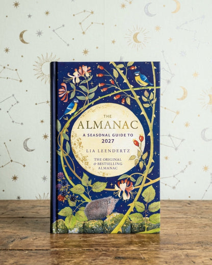 The Almanac: A Seasonal Guide to 2027 by Lia Leendertsz, a hardcover book with a dark blue cover decorated with whimsical botanical and celestial illustrations, standing on a rustic wooden table against a patterned wallpaper background.