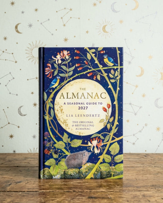 The Almanac: A Seasonal Guide to 2027 by Lia Leendertsz, a hardcover book with a dark blue cover decorated with whimsical botanical and celestial illustrations, standing on a rustic wooden table against a patterned wallpaper background.