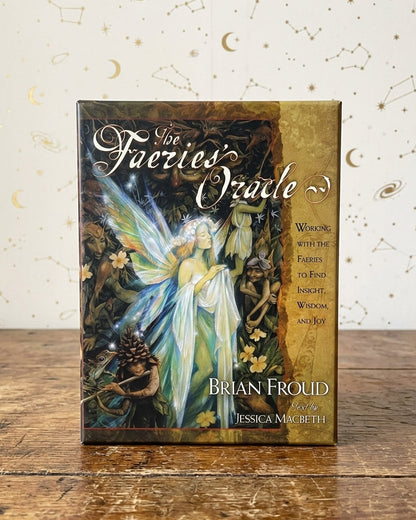 The box of 'The Faeries Oracle' by Brian Frouid stands on a rustic wooden table against a cream-colored background patterned with gold stars and constellations.