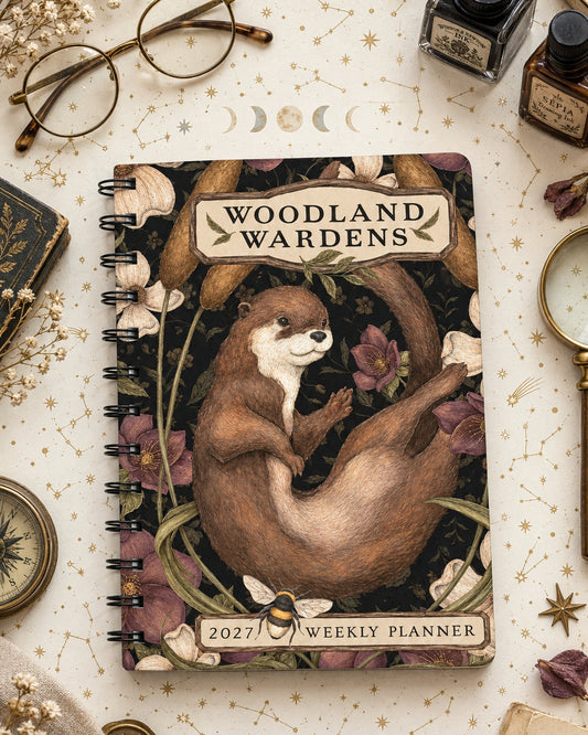 A spiral-bound 2027 weekly planner titled 'Woodland Wardens' featuring an illustration of an otter among flowers and leaves on a dark background, styled on a light patterned surface.