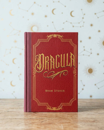 A red hardcover book titled 'Dracula' by Bram Stoker, standing upright on a wooden surface against a light blue background decorated with gold celestial patterns.