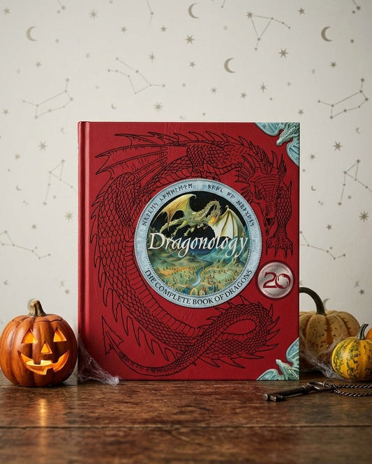 A red hardcover book titled "Dragonology: The Complete Book of Dragons" stands on a wooden surface against a light wall with moon and constellation illustrations. Halloween-themed pumpkins are placed around it as props.