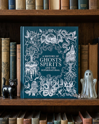 A hardcover book titled "A History of Ghosts Spirits and the Supernatural" by DK stands upright on a wooden bookshelf, surrounded by vintage books. The cover is dark teal with intricate white gothic illustrations of a ghost, tree, roots, and other spirits.