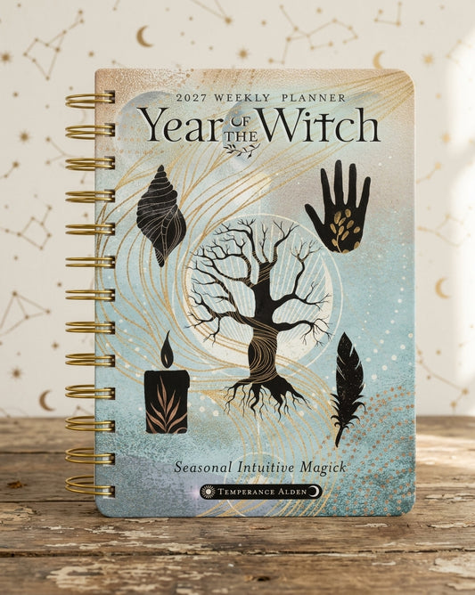 A spiral-bound 2027 Weekly Planner titled 'Year of the Witch' by Temperance Alden, featuring a mystical design with a tree, candles, feathers, and a shell on a light blue and beige background, displayed on a rustic wooden surface.