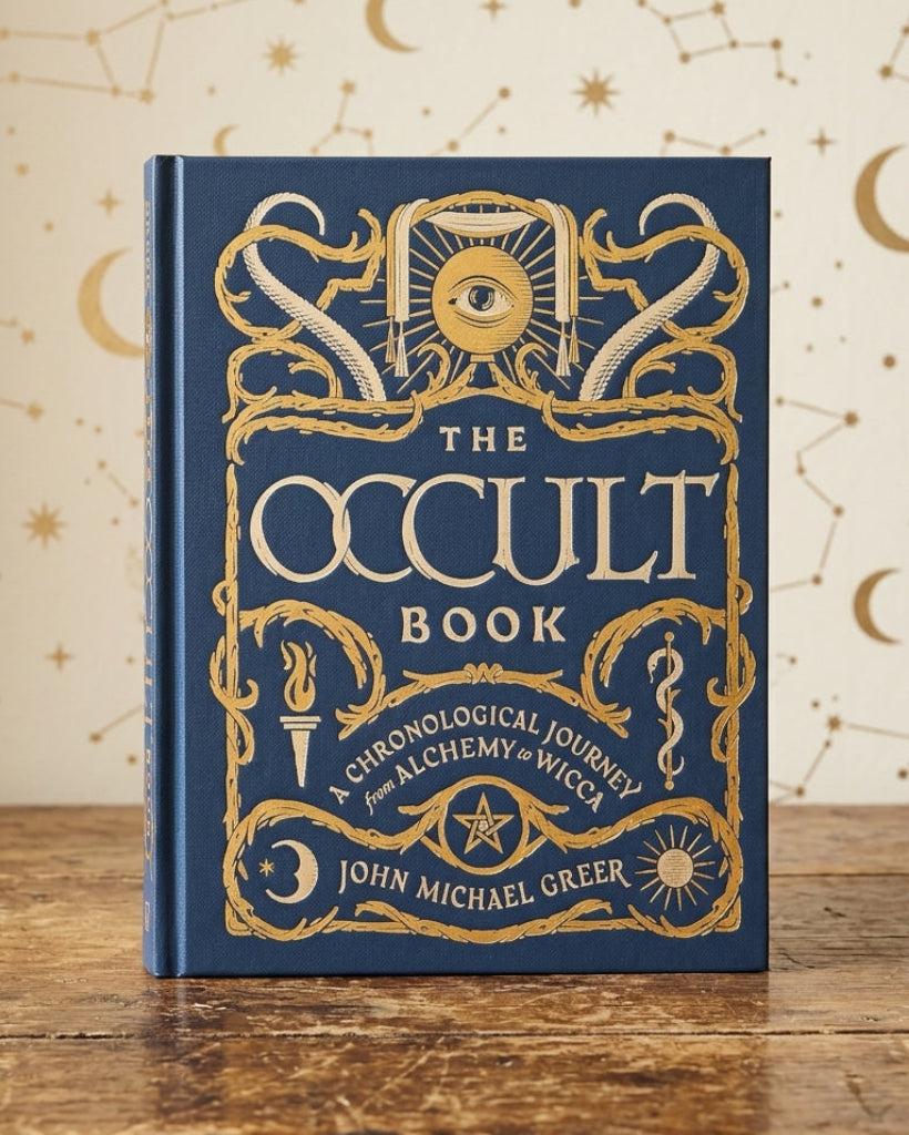 The Occult Book by John Michael Greer, a hardcover book with a deep blue cover and ornate gold foil illustrations, standing on a rustic wooden table against a cream-colored background decorated with gold celestial patterns.