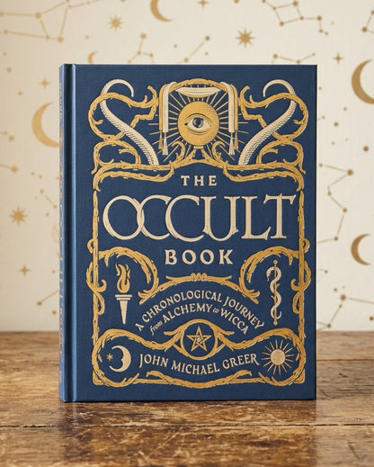 The Occult Book by John Michael Greer, a hardcover book with a deep blue cover and ornate gold foil illustrations, standing on a rustic wooden table against a cream-colored background decorated with gold celestial patterns.