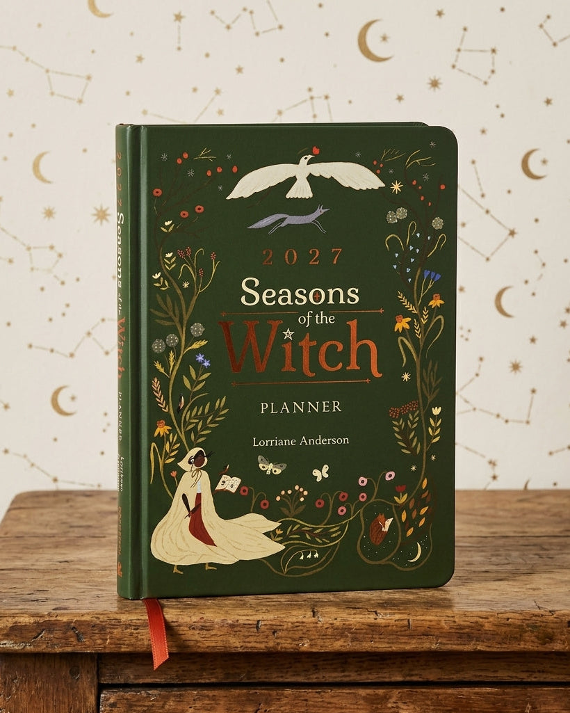 A hardcover planner titled '2027 Seasons of the Witch Planner' by Lorriane Anderson, featuring a dark green cover decorated with whimsical witch illustrations and gold lettering, displayed on a rustic wooden table against a cream wall with celestial patterns.