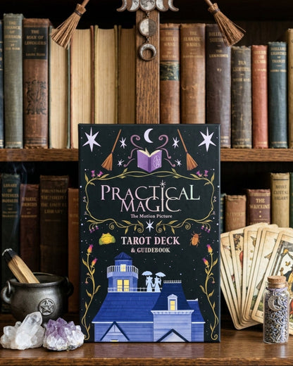 The book 'Practical Magic Tarot Deck & Guidebook' stands upright on a wooden shelf in front of a bookcase filled with old books. A small cauldron, crystals, a card deck, and a jar are arranged around it.