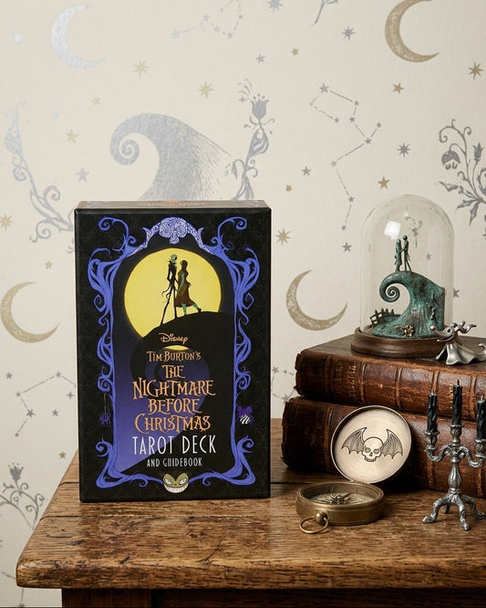 The Burton's The Nightmare Before Christmas Tarot Deck and Card Book box displayed on a rustic wooden table next to themed decor, including a glass dome, candles, and a compass, against a patterned wallpaper background.