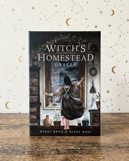 The book 'Witch's Homestead Oracle' by Hadas Knox and Giada Rose is displayed standing upright on a rustic wooden table against a cream-colored background decorated with gold crescent moons and constellations.