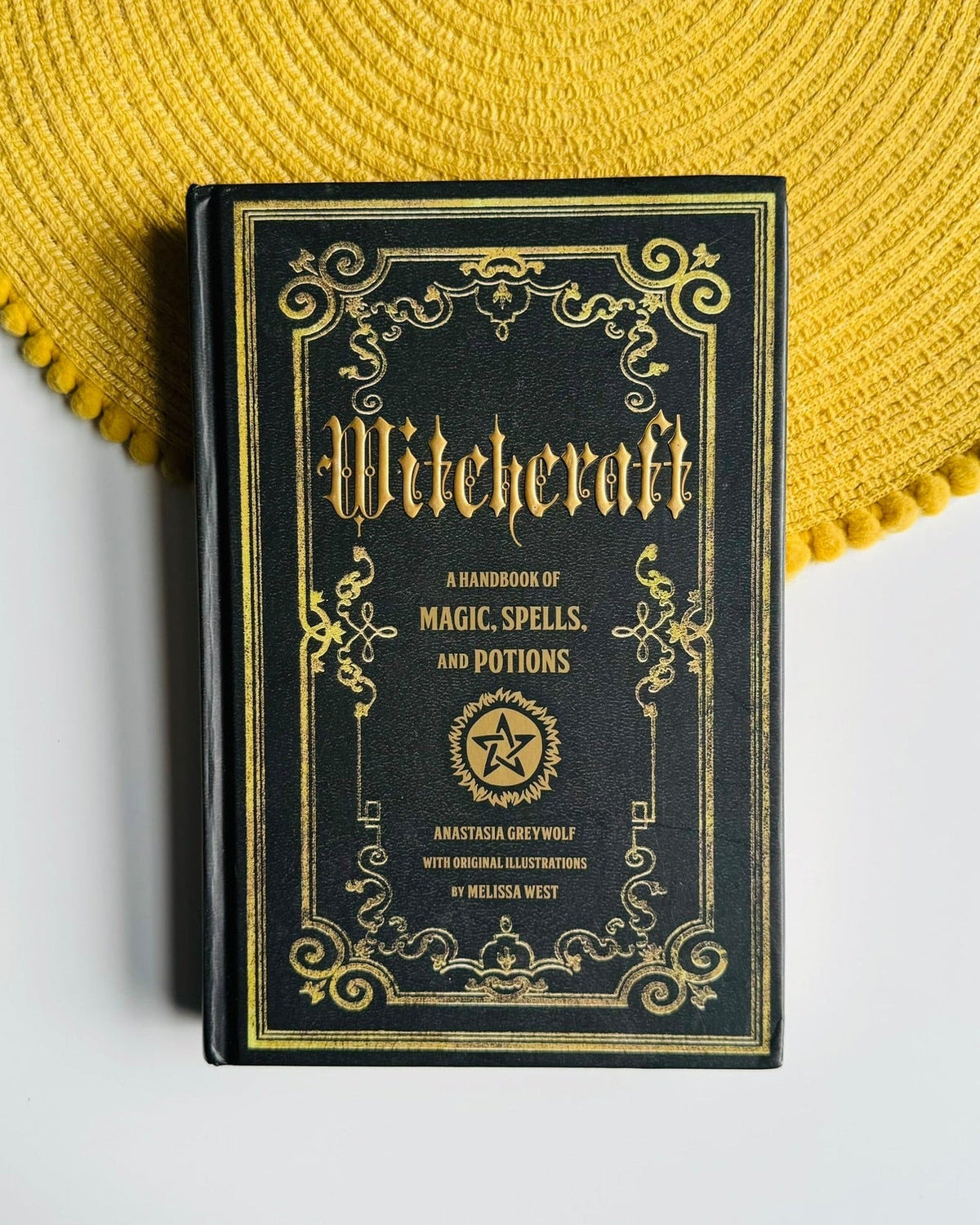A black hardcover book titled 'Witchcraft: A Handbook of Magic, Spells, and Potions' with ornate gold detailing, styled on a white surface next to a yellow woven placemat.