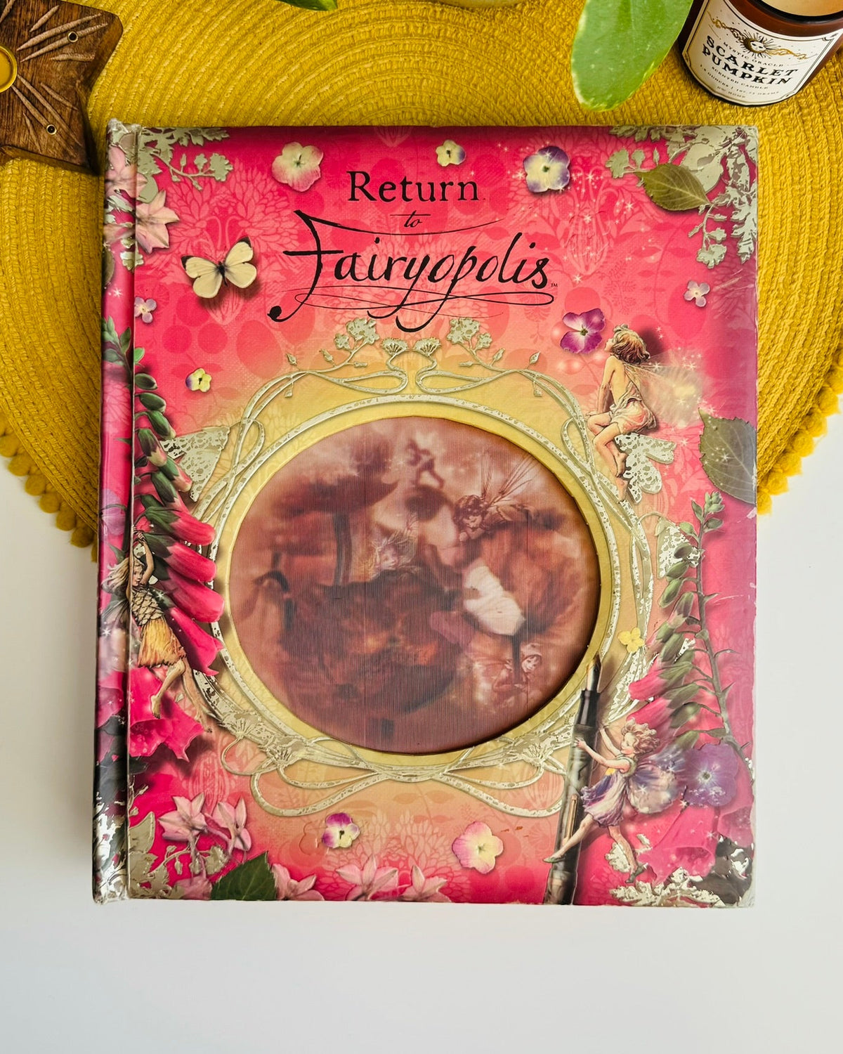 A decorative hardcover book titled "Return to the Fairypolis" with a pink floral cover and a circular cutout showing a vintage fairy illustration, styled on a yellow woven placemat with a bottle partially visible at the top right.