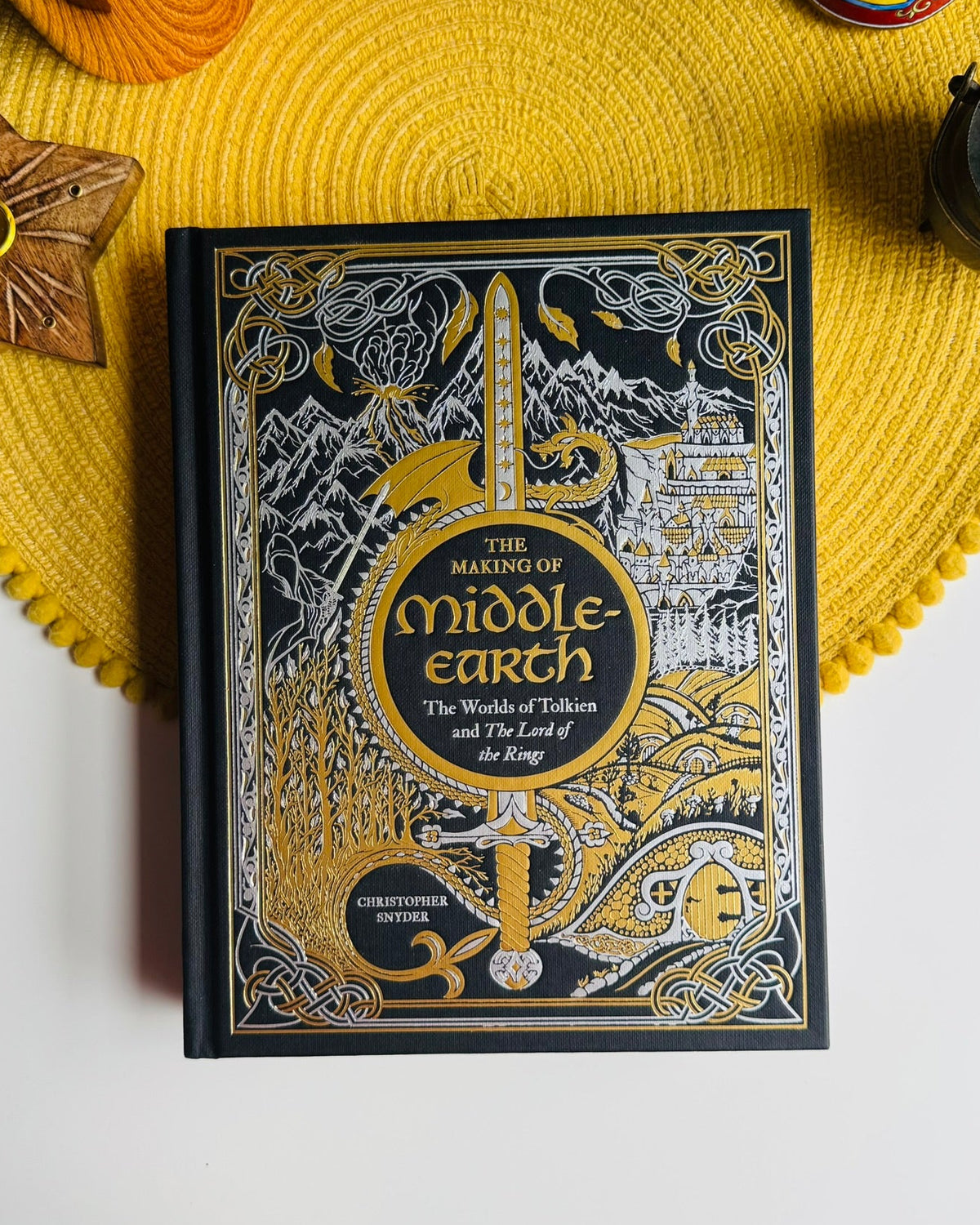 The Making of Middle-earth: The Worlds of Tolkien and The Lord of the Rings book by Christopher Fowler, styled on a yellow woven placemat with decorative props.