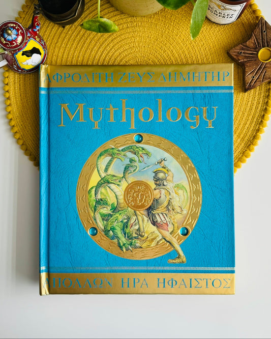 A hardcover book titled "Mythology" with a bright blue cover and gold lettering, placed on a yellow woven placemat with decorative props around it.