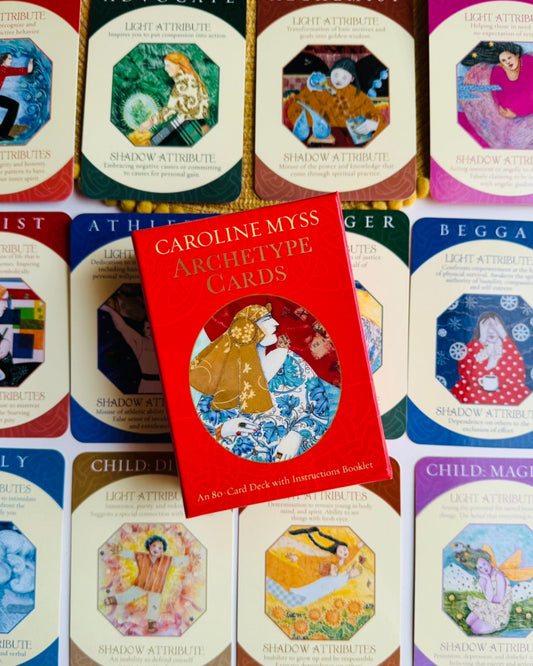 A set of colorful tarot-style cards arranged in rows, with a red book titled 'Caroline Myss Algebrite Cards' placed in the center. The cards feature various illustrated faces and symbols on colorful backgrounds.