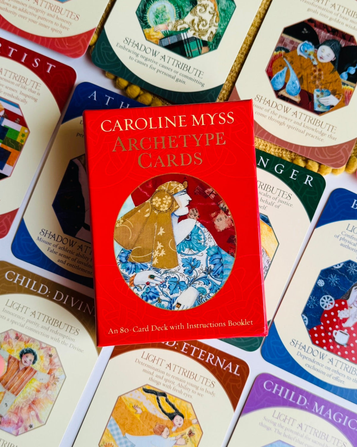 A red box of Caroline Myss Archetype Cards is centered on a spread of matching illustrated cards, with a hand visible at the lower left edge.