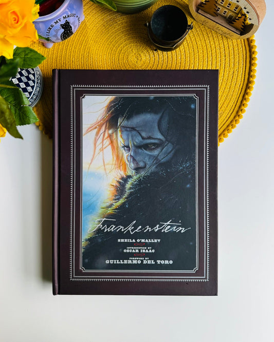 A hardcover book titled 'Frankenstein' by Sheila O'Malley, Series Tales, Guillamete Sel Varga, styled on a yellow woven placemat with flowers and other decorative items around it.