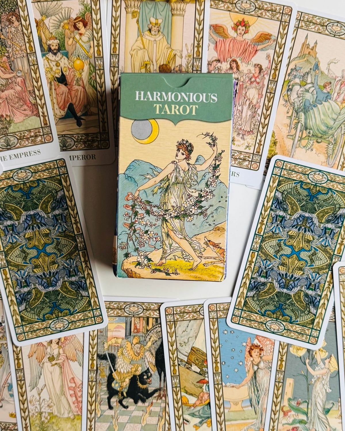 A box of Harmonious Tarot cards is centered among several illustrated tarot cards spread out on a white surface. The box has a green top band and vintage-style artwork of a woman holding flowers.