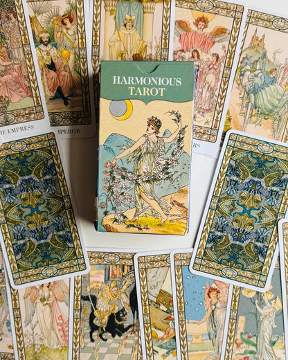 A box of Harmonious Tarot cards is centered among several illustrated tarot cards spread out on a white surface. The box has a green top band and vintage-style artwork of a woman holding flowers.