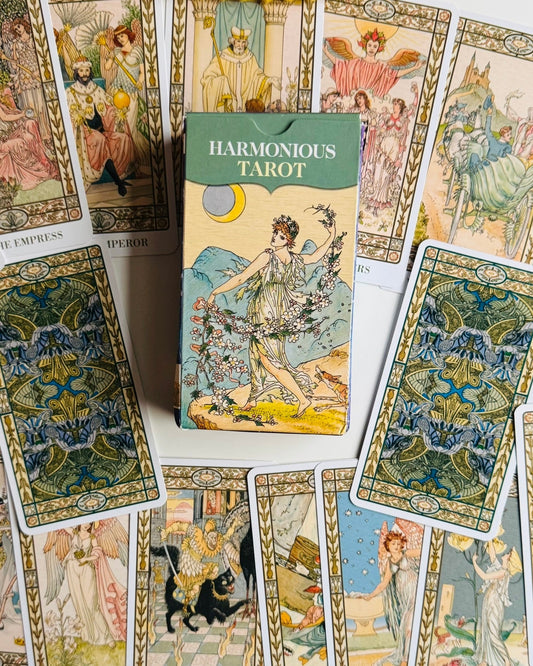 A box of Harmonious Tarot cards is centered among several illustrated tarot cards spread out on a white surface. The box has a green top band and vintage-style artwork of a woman holding flowers.