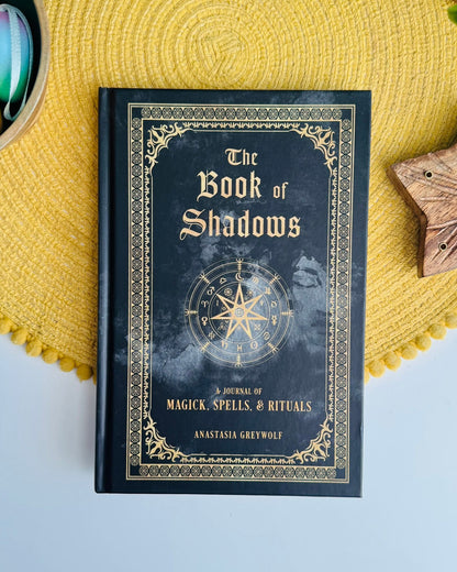 A black hardcover book titled 'The Book of Shadows' with gold gothic lettering and an ornate gold border, placed on a yellow woven placemat. The cover features a compass rose illustration and the subtitle 'A Journal of Magick, Spells & Rituals' by Anastasia Greywolf.