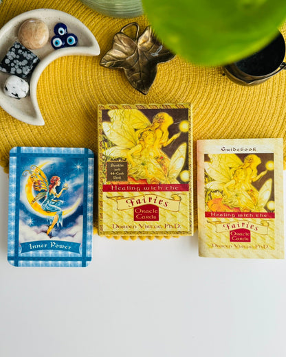 Three Fairies Oracle Cards products arranged on a yellow woven mat: an Inner Power tarot card deck, a Healing with Me oracle card deck box, and a Quirksbook Healing with Me oracle card deck box, styled with crystals, a small dish, and a green leaf in the background.