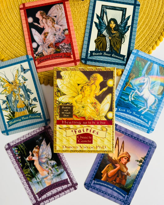 A set of illustrated fairy-themed tarot cards arranged on a yellow woven placemat over a white surface. The central card reads 'Healing with the Fairies' and is signed by Dr.