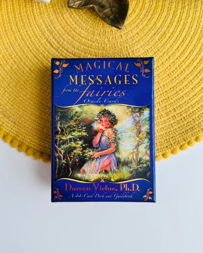 A boxed card deck titled "Magical Messages from the Fairies" lies on a yellow woven placemat. The box is dark blue with gold lettering and an illustration of a fairy in a forest.