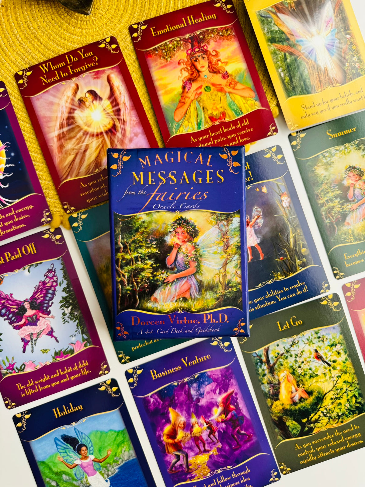 A spread of colorful oracle or card deck boxes and cards featuring fairy illustrations and titles such as 'Magical Messages from the Fairies,' 'Emotional Healing,' 'Business Venture,' and 'Holiday.' The products are arranged on a light surface with a yellow woven textile underneath.