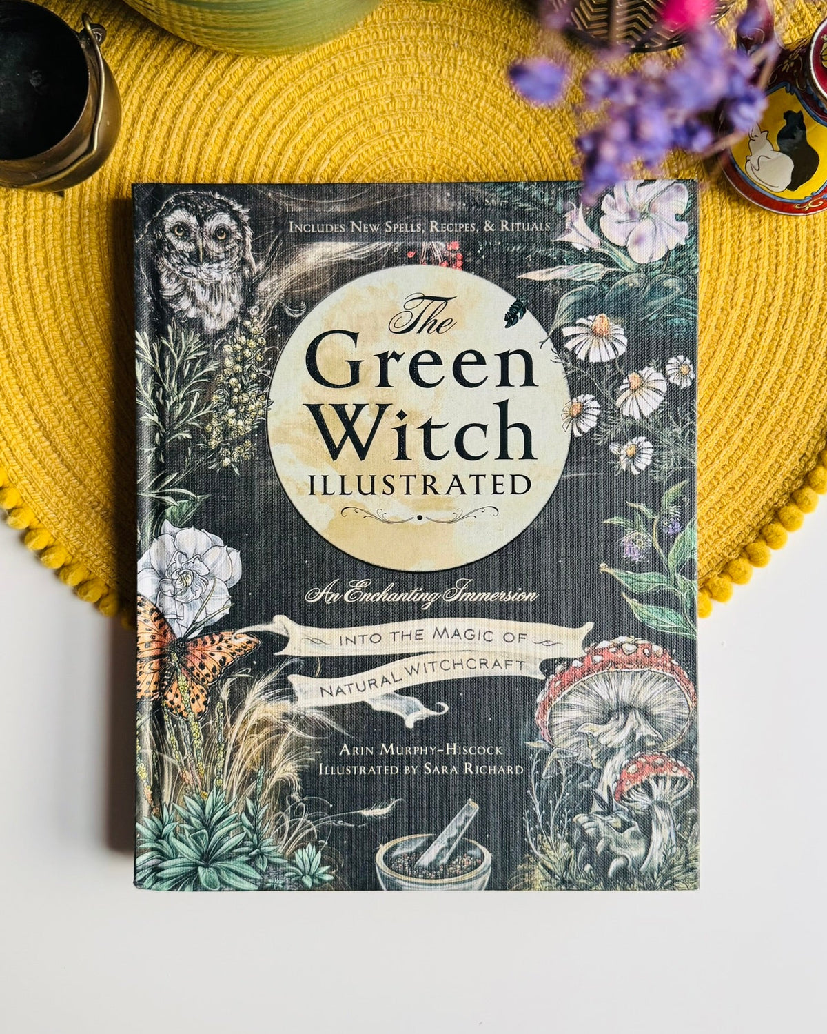 The Green Witch Illustrated book by Asin Murphy-Hieuce and Sarah Richard, featuring an illustrated cover with a dark background and botanical designs, styled on a yellow woven placemat with decorative props.