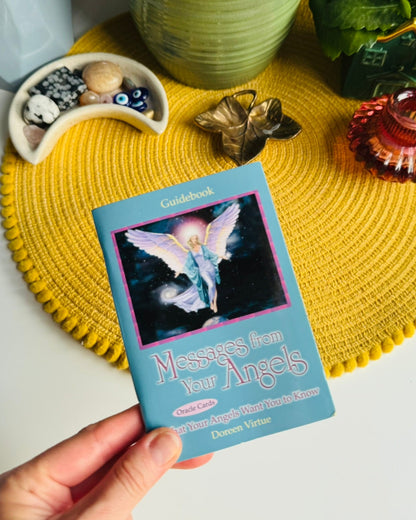 A hand holds a blue card deck box titled "Messages from Angels" in front of a yellow woven placemat on a white table, with potted plants and small decorative objects in the background.
