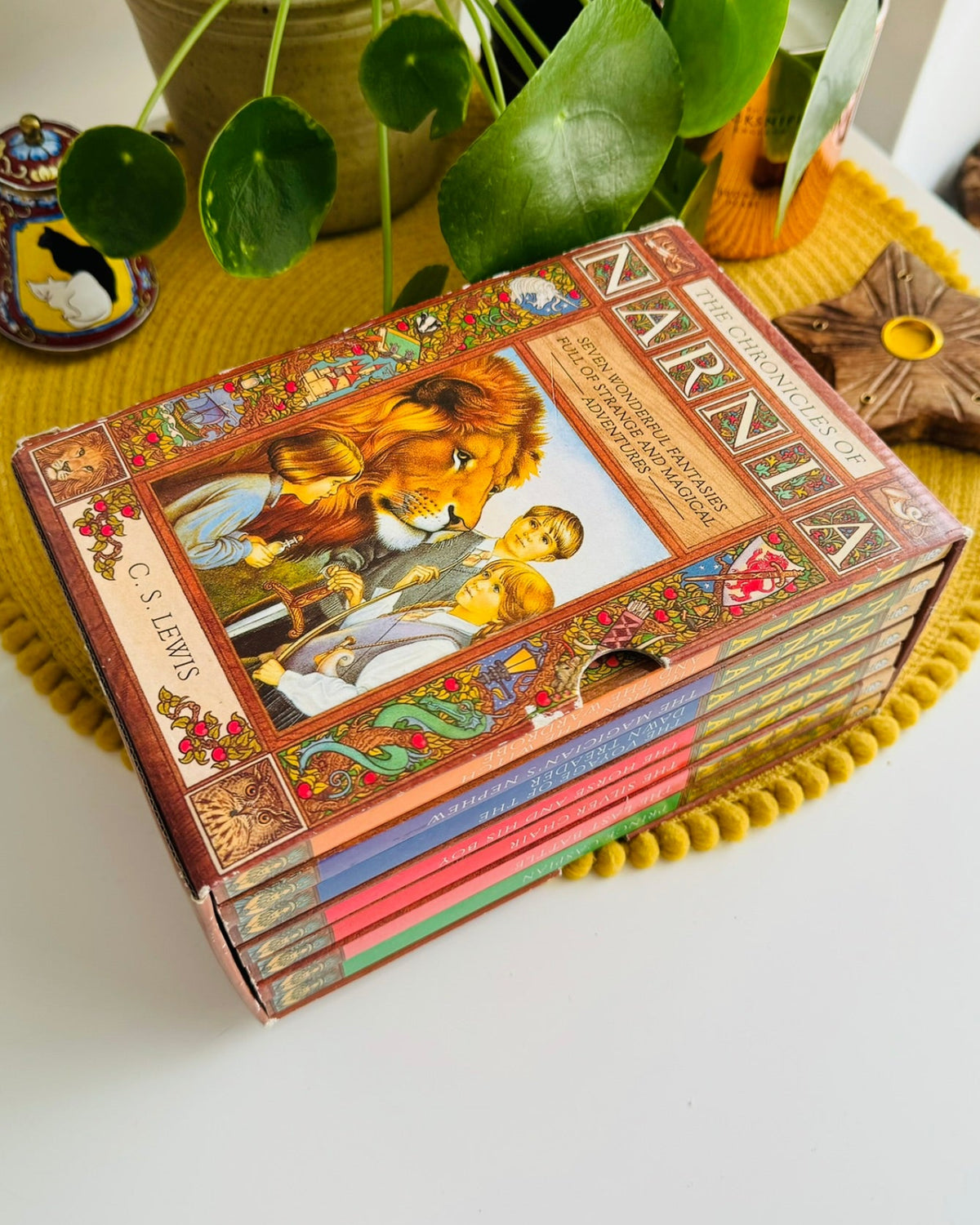 A boxed set of children's books titled 'NARNA' with illustrated covers, arranged on a yellow pom-pom placemat on a white table with potted plants and decorative items in the background.