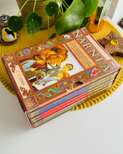 A boxed set of children's books titled 'NARNA' with illustrated covers, arranged on a yellow pom-pom placemat on a white table with potted plants and decorative items in the background.